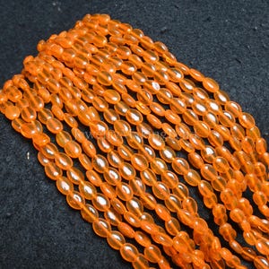 Può includere: Una collezione di perline ovali traslucide di colore arancione. Le perline sono infilate insieme, creando più fili. Le perline hanno una finitura lucida e sono disposte diagonalmente su una superficie scura.