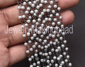 Op de afbeelding: Een streng witte parels die aan een zwarte ketting zijn geregen. De ketting is gemaakt van kleine, met elkaar verbonden metalen ringen. De parels zijn gelijkmatig verdeeld en de ketting is doorlopend.