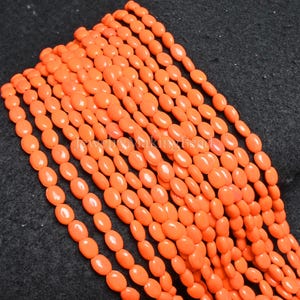 Puede incluir: Primer plano de múltiples hebras de cuentas ovaladas naranjas. Las cuentas son de tamaño y forma uniformes, con un acabado brillante. Las cuentas están dispuestas en líneas paralelas, creando una superficie texturizada. El fondo es de color oscuro.