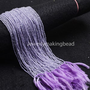 May include: A strand of light purple amethyst beads, approximately 2mm in diameter, with a tassel at the end. The beads are strung on a clear thread and are arranged in a long, flowing strand. The strand is lying on a black surface.
