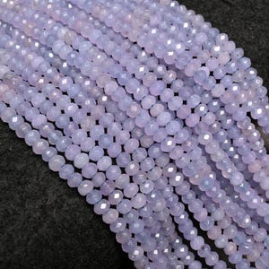 May include: Close-up of multiple strands of light purple, faceted beads. The beads are small and round, with a subtle sheen. They are strung together, creating a textured effect. The background is a dark grey.