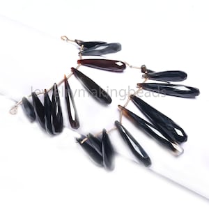 Könnte beinhalten: Stränge aus facettierten, tropfenförmigen schwarzen Edelsteinen mit goldfarbenem Draht. Die Edelsteine haben eine polierte, reflektierende Oberfläche und variieren in der Größe. Die Anordnung wird auf einer weißen Oberfläche gezeigt.