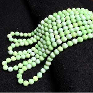 May include: A strand of light green gemstone beads. The beads are round and smooth, and they are strung together on a clear thread. The beads are arranged in a loose pile, and they are illuminated by a soft light.