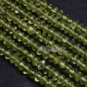 Puede incluir: Primer plano de hilos de cuentas de piedras preciosas de peridoto verde oliva. Las cuentas tienen una forma orgánica única, con un centro redondeado y extremos acampanados, ensartadas juntas. Las cuentas son translúcidas y tienen un acabado pulido.