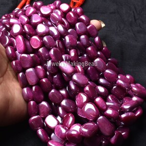 May include: Strand of polished, oval-shaped ruby beads. The beads are a deep, rich red-purple color with a glossy finish. The beads are strung together with a red cord. The beads are approximately 0.5 inches in length.
