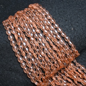 Puede incluir: Primer plano de hebras de cuentas ovales de color melocotón. Las cuentas translúcidas están ensartadas, creando una forma fluida. Las cuentas parecen ser de vidrio o un material similar. El fondo es una tela oscura y texturizada.