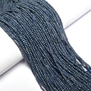 Puede incluir: Primer plano de múltiples hebras de pequeñas cuentas facetadas de color azul grisáceo. Las cuentas están ensartadas, creando una apariencia fluida y texturizada. Las cuentas tienen un brillo sutil. Ideal para la fabricación de joyas.