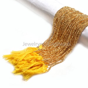 May include: Close-up of gold-colored beaded chains with yellow tassels. The chains are draped over a white surface. The beads have a faceted texture, reflecting light. The tassels are made of yellow thread.