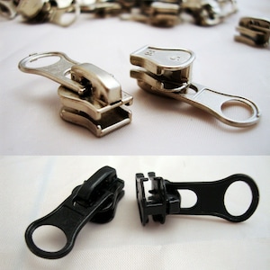 May include: Two sets of zipper pulls, one silver and one black. The silver zipper pull has a loop on the end and a slider with a small metal tab. The black zipper pull has a loop on the end and a slider with a small black tab.