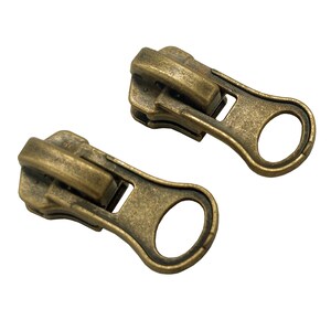 May include: Two antique gold zipper pulls with a loop on the end. The zipper pulls are made of metal and have a worn, aged look.
