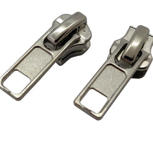 May include: Two silver zipper pulls with a rectangular shape and a square opening in the center. The pulls have a smooth, metallic finish.