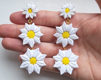 May include: A pair of white daisy earrings with yellow centers. Each earring has three daisies stacked on top of each other.
