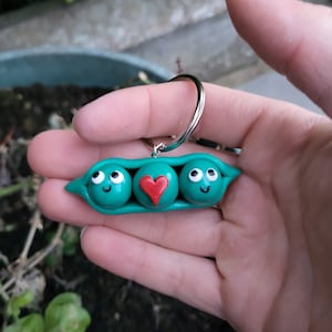 May include: A green pea pod keychain with three peas. The middle pea has a red heart on it, and the other two peas have smiling faces.