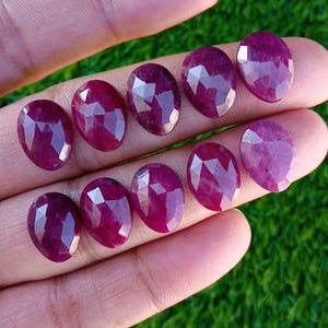 May include: Ten oval-cut rubies with multiple facets, displayed on a hand. The gemstones are a deep red-purple colour, reflecting light across their polished surfaces. Each ruby is a unique shape, showcasing the natural beauty of the stones.