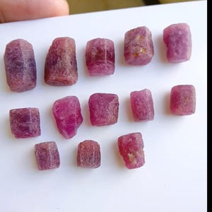 Puede incluir: Colección de piedras preciosas de rubí en bruto. Las piedras son de un color rojo rosado intenso, con una superficie rugosa y texturizada. Las piedras son de forma rectangular y cilíndrica, y varían ligeramente en tamaño.