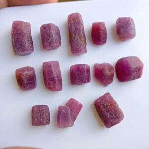 Peut inclure: Une collection de pierres précieuses rubis brutes et non taillées. Les pierres, de tailles et de formes variées, présentent une teinte rouge-violet foncé. La texture rugueuse et les formes naturelles des rubis sont visibles.