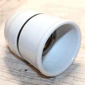 Puede incluir: Un portalámparas de cerámica blanca con una banda negra en el centro. El portalámparas cilíndrico tiene una abertura ancha y está sobre papel impreso. El interior del portalámparas es visible.