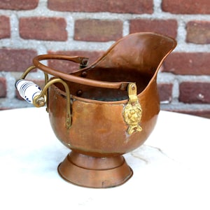 May include: A copper coal scuttle with a handle and a decorative brass lion emblem. The handle has a white and blue ceramic grip. The scuttle is set against a brick wall and a white surface.
