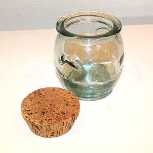 May include: A clear glass jar with a rounded body and a wide opening, alongside a cork stopper. The jar has a slight green tint and two small handles. The cork is a light brown colour. The items are on a white surface.