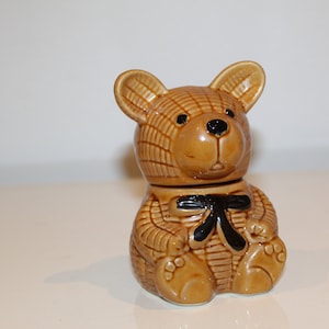 Puede incluir: Un tarro de miel de cerámica con forma de oso de peluche marrón con una pajarita negra. El oso tiene una superficie texturizada y ojos negros.