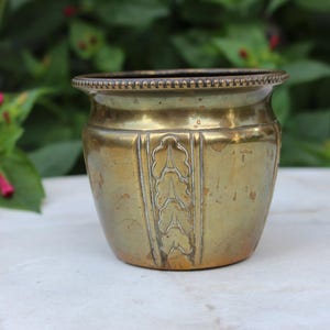 Op de afbeelding: Een kleine, antieke messing plantenbak met een decoratief bladontwerp. De plantenbak heeft een afgeronde body en een licht uitlopende rand met een kralenrand. Het messing heeft een warme, verouderde patina, wat wijst op de vintage oorsprong.