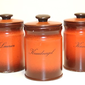 Op de afbeelding: Drie keramische kruidenpotten met deksels. De potten zijn oranje met een bruine basis en deksel. De potten hebben tekst, waaronder "Laurier", "Kruidnagel" en "Kaneel".