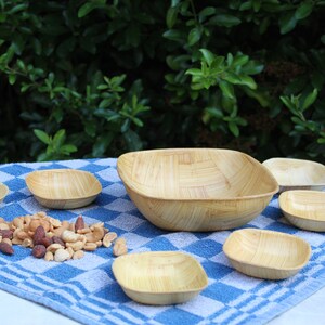 Peut inclure: Un ensemble de sept bols en bois, comprenant un grand bol carré et six petits bols ronds, disposés sur une nappe à carreaux bleus et blancs. Les bols sont en bambou naturel et sont parfaits pour servir des en-cas ou des amuse-gueules.