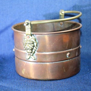 Peut inclure: Seau en cuivre avec une poignée en laiton et des détails de tête de lion. Le seau cylindrique comporte deux bandes horizontales et des rivets. La poignée arquée et la tête de lion ajoutent des éléments décoratifs.