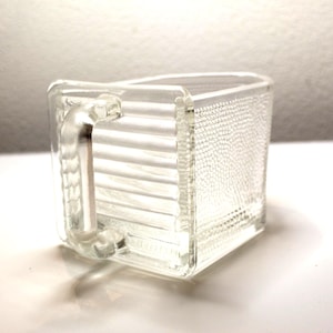 May include: A clear, rectangular glass mug with a handle. The mug's side has a textured, dotted pattern, while the front has horizontal lines. The handle is also clear glass. The mug is sitting on a white surface.