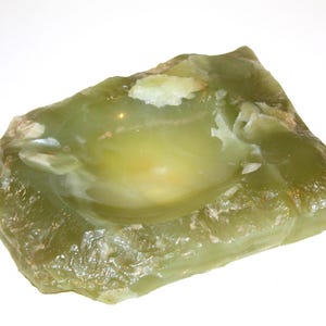 May include: A roughly-hewn, olive-green stone bowl with a smooth, rounded interior. The natural stone has a rough, textured exterior and a lighter, yellowish-green center. The bowl is set against a white background.