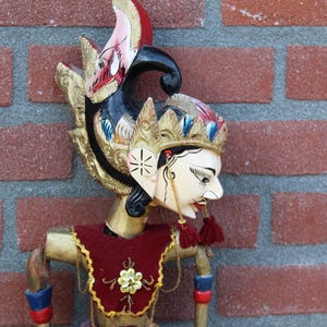 Puede incluir: Un títere de madera con un tocado colorido y una cara pintada. El títere lleva un traje rojo y dorado y tiene un tocado largo y fluido.