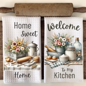 May include: Two white kitchen towels with a farmhouse theme. The first towel has a floral arrangement, a rolling pin, a pot, and a jar of cookies with the text "Home Sweet Home". The second towel has a floral arrangement, a rolling pin, a bowl of cookies, a jar, and a bottle with the text "Welcome To My Kitchen".