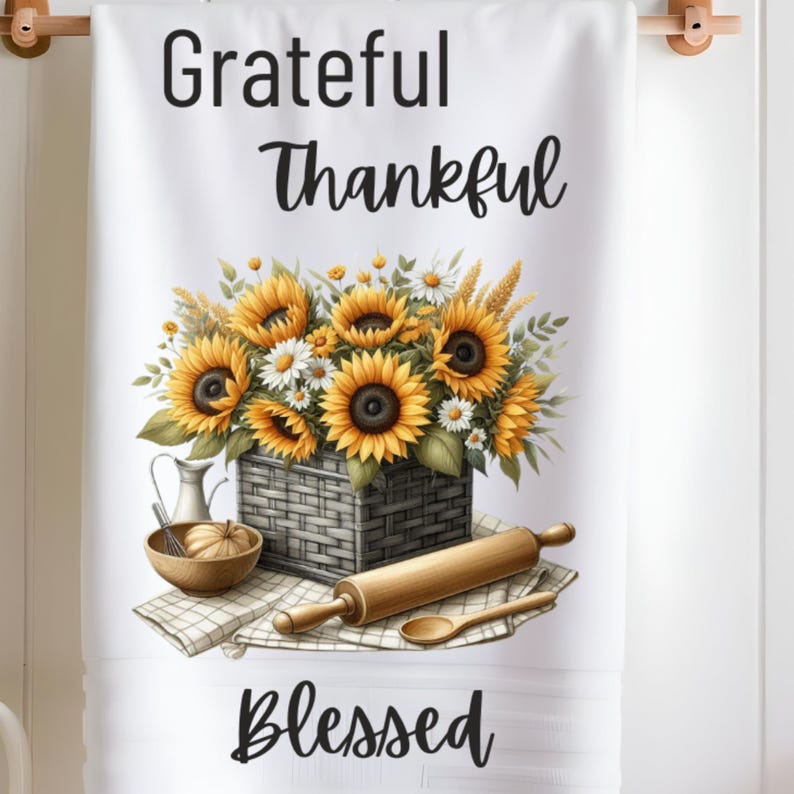 Puede incluir: Toalla de cocina blanca con un dise&ntilde;o floral de girasoles y margaritas blancas en una cesta tejida. El texto "Grateful Thankful Blessed" est&aacute; impreso en la toalla.