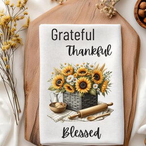 Puede incluir: Una toalla de cocina blanca con un dise&ntilde;o floral con girasoles y margaritas en una cesta. El texto "Grateful Thankful Blessed" est&aacute; impreso en la toalla.