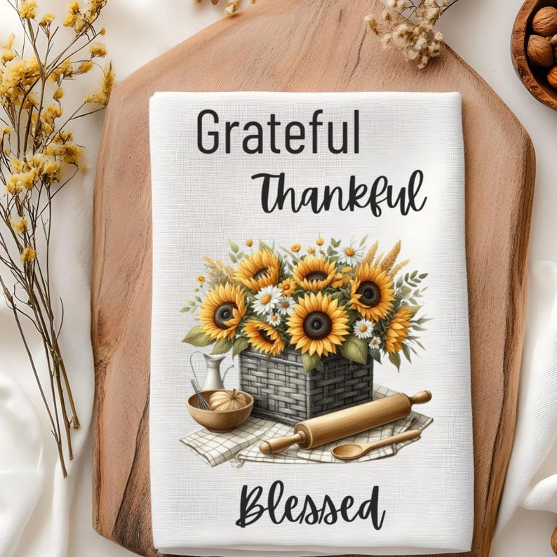 Puede incluir: Una toalla de cocina blanca con un dise&ntilde;o floral con girasoles y margaritas en una cesta. El texto "Grateful Thankful Blessed" est&aacute; impreso en la toalla.