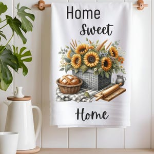 Puede incluir: Toalla de cocina blanca con el texto "Home Sweet Home" impreso en negro. La toalla presenta una ilustraci&oacute;n de acuarela de una cesta de girasoles, una hogaza de pan en una cesta y un rodillo.