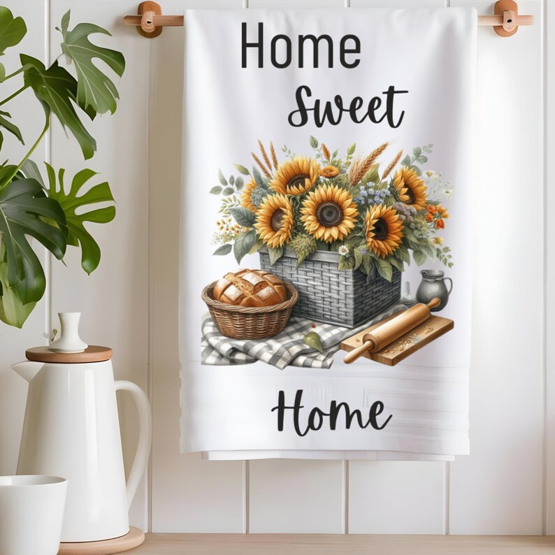 Puede incluir: Toalla de cocina blanca con el texto "Home Sweet Home" impreso en negro. La toalla presenta una ilustraci&oacute;n de acuarela de una cesta de girasoles, una hogaza de pan en una cesta y un rodillo.