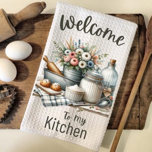 May include: White kitchen towel with a floral design and the text "Welcome to my Kitchen". The towel features a vintage-style illustration of a kitchen scene with a rolling pin, a jar of flour, a bowl of sugar, and a bouquet of flowers.