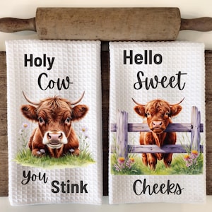 Puede incluir: Dos toallas de cocina blancas con un diseño de vaca Highland marrón y blanca. La primera toalla dice "Holy Cow You Stink" y la segunda toalla dice "Hello Sweet Cheeks".