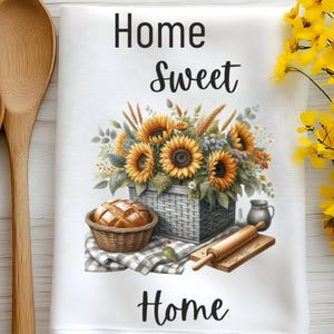 Puede incluir: Una toalla de cocina blanca con el texto "Home Sweet Home" impreso en negro. La toalla presenta una cesta de girasoles, una hogaza de pan en una cesta de mimbre, un rodillo y una jarra.