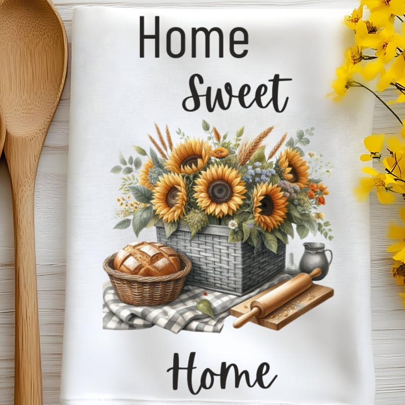 Puede incluir: Una toalla de cocina blanca con el texto "Home Sweet Home" impreso en negro. La toalla presenta una cesta de girasoles, una hogaza de pan en una cesta de mimbre, un rodillo y una jarra.