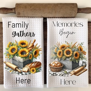 May include: Two white kitchen towels with a waffle weave texture. The left towel features a basket of sunflowers with the text "Family gathers here". The right towel features a basket of sunflowers with the text "Memories begin here".