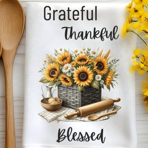 Puede incluir: Toalla de cocina blanca con un ramo de girasoles en una cesta, un rodillo y una cuchara de madera. El texto "Grateful Thankful Blessed" est&aacute; impreso en la toalla.