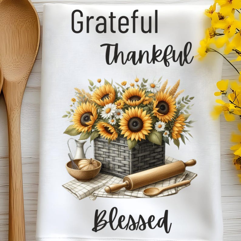 Puede incluir: Toalla de cocina blanca con un ramo de girasoles en una cesta, un rodillo y una cuchara de madera. El texto "Grateful Thankful Blessed" est&aacute; impreso en la toalla.
