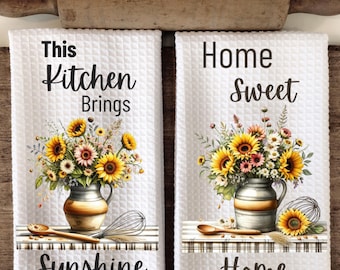 May include: Two white kitchen towels with sunflower designs and text. The first towel says "This Kitchen Brings Sunshine" and the second towel says "Home Sweet Home".