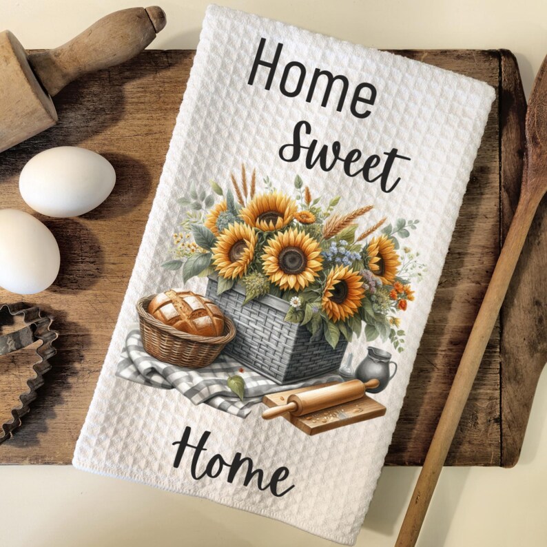 Puede incluir: Toalla de cocina blanca con textura de waffle. La toalla presenta una cesta de girasoles, una hogaza de pan y un rodillo. El texto "Home Sweet Home" est&aacute; impreso en la toalla.
