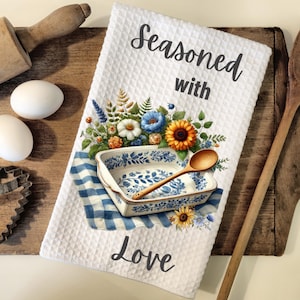 May include: White kitchen towel with a blue and white patterned baking dish, a wooden spoon, and a bouquet of flowers. The text "Seasoned with Love" is printed on the towel.