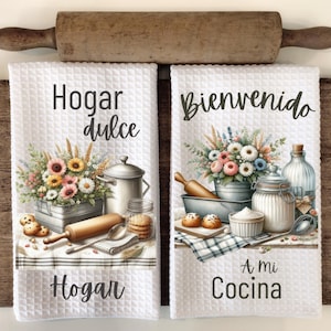 May include: Two white kitchen towels with a waffle weave texture. The left towel features a floral arrangement with a rolling pin, a spoon, and a jar of cookies. The text on the towel reads "Hogar dulce Hogar". The right towel features a floral arrangement with a rolling pin, a loaf pan, a jar of sugar, a bottle, and a bowl. The text on the towel reads "Bienvenido a mi Cocina".