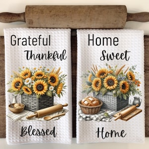 Puede incluir: Dos toallas de cocina blancas con dise&ntilde;os de girasoles y trigo. La primera toalla dice "Grateful Thankful Blessed" y la segunda toalla dice "Home Sweet Home".