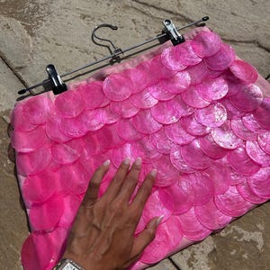 Peut inclure: Une jupe rose ornée d'un motif en écailles superposées composé de disques circulaires, translucides et iridescents, fixés sur une base en tissu. Le vêtement est suspendu à un cintre métallique muni de deux pinces noires sur la ceinture.